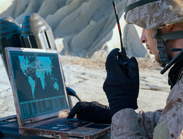Soldier talking on a handheld satellite radio, looking at a map on a laptop