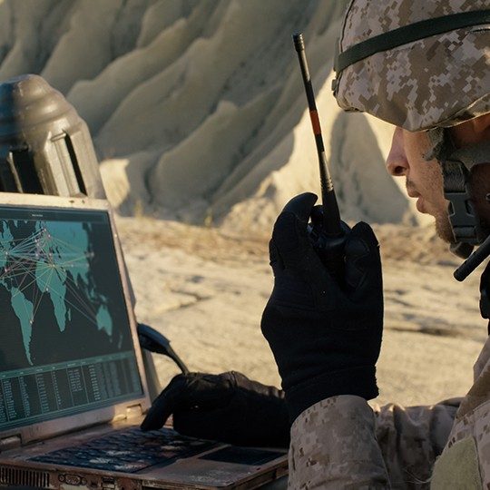 Man dressed in combat gear talking on a radio while looking at a map of the world on a miltary laptop