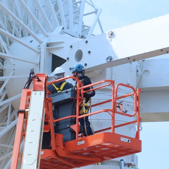 Two men in hardhats standing in an aerial work platform doing antenna maintenance on the back of a ground station