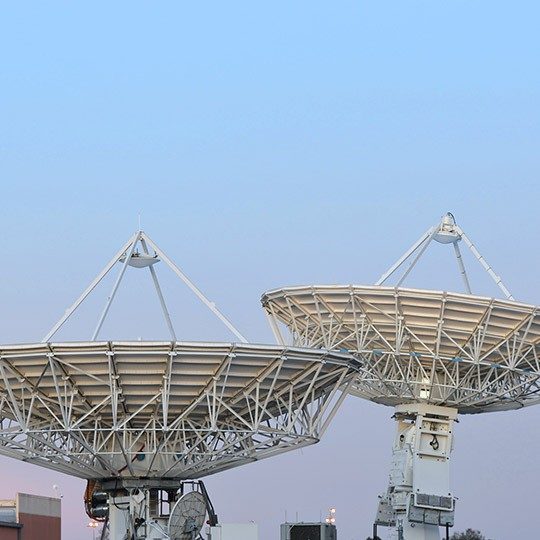 Two large, white gateway SATCOM antennas pointed at a light blue sky at dusk