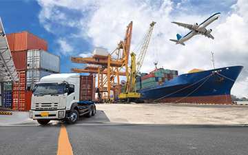 Truck parked on a dock next to cargo containers and a cargo ship with an airplane flying above