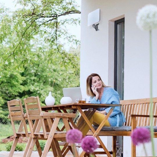 Woman sitting outside of her home at a wood table talking on the phone in front of a laptop