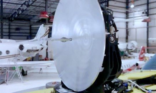 Parabolic reflector antenna on a table inside a hanger with an airplane in the background