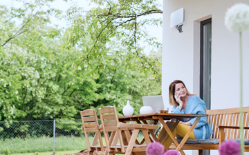 Woman sitting outside of her home at a wood table talking on the phone in front of a laptop