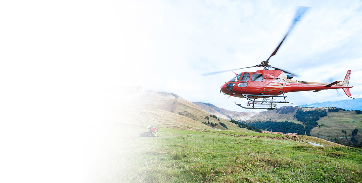 Red helicopter flying over a man kneeled down in the grass on the side of a mountain