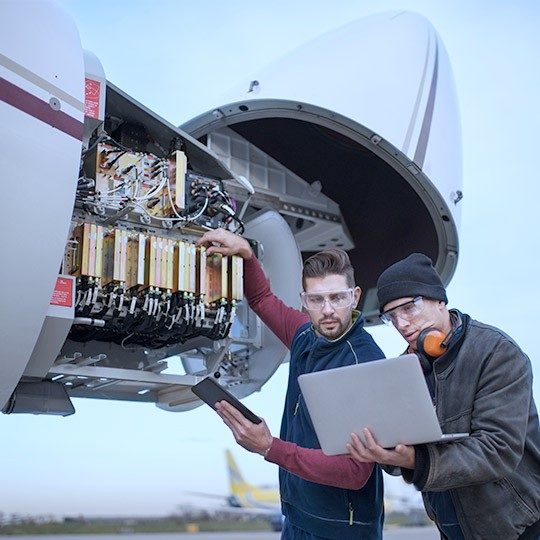 Two male plane technicians working on the nose of a plane, reviewing informaiton on a laptop