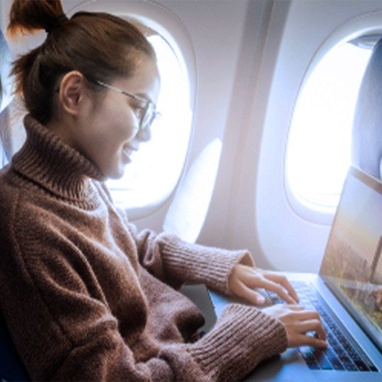 Woman wearing glasses and a brown sweater is working during a flight on her laptop over onbaord WiFi