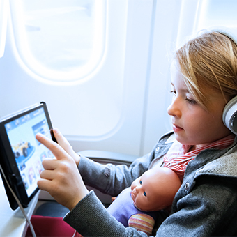 Young girl wearing headphones with a baby doll in her lap playing a game on her tablet over inflight WiFi