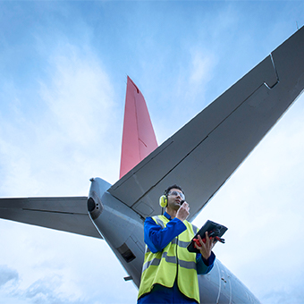 An aiport employee in a bright yellow fest standing under the tail wings of a plane holding a tablet