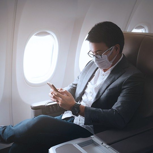 Male passenger wearing a business suit and face mask using airplane satellite internet on his smart phone