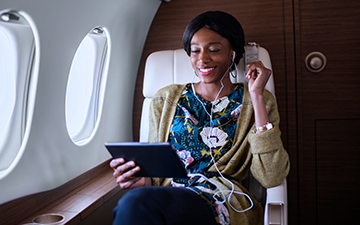 Woman with black hair and a floral shirt flying in a private jet, wearing a headset connected to her tablet