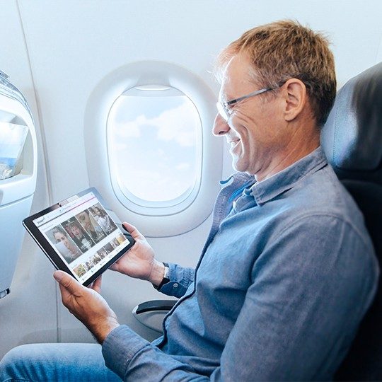 A smiling man wearing glasses is holding a tablet viewing inflight marketing content on his tablet