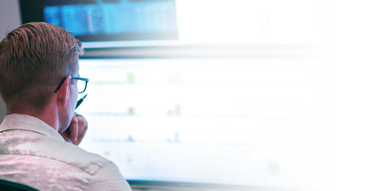 Blonde man in glasses holding a pen, staring at a computer monitor displaying information connected by franchise internet