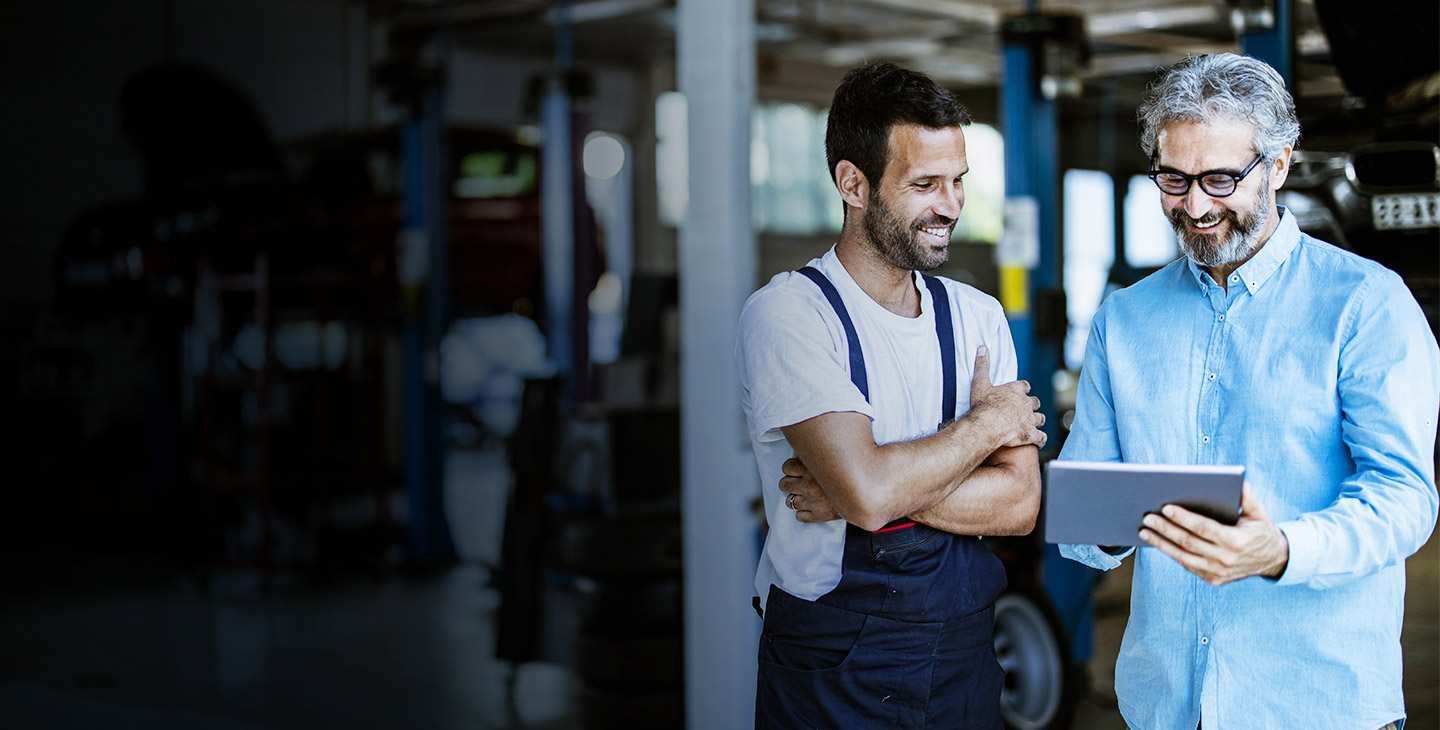 Two men in an auto repair shop smiling, communicating with Viasat business customer service on a laptop