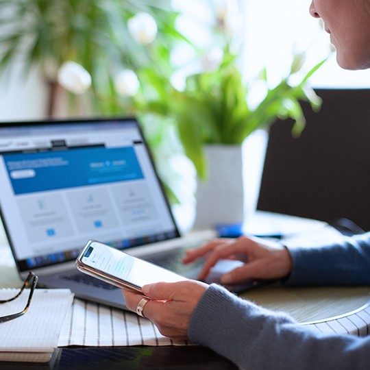 Professional woman sitting at a desk with a cell phone in-hand, managing her Viasat business account on a laptop