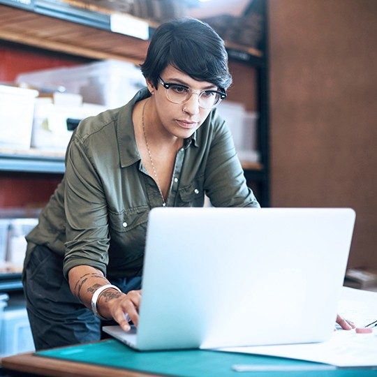 Dark-haired woman in forrest green shirt on a laptop in a storage room