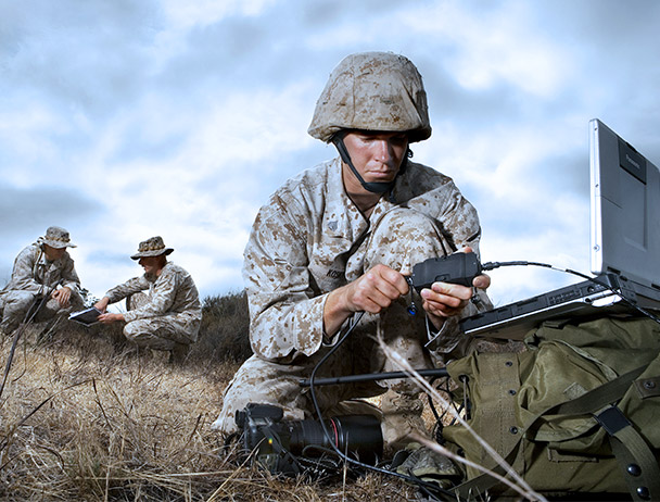 Three military soldiers in kneeling in the field, wearing camo and working on communications equipment