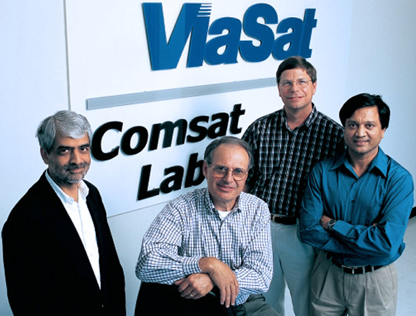 Four Viasat employees smiling at the camera in front of a Viasat Comsat Lab sign