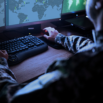 Military man sitting at a desktop computer looking at a map of the world on a monitor