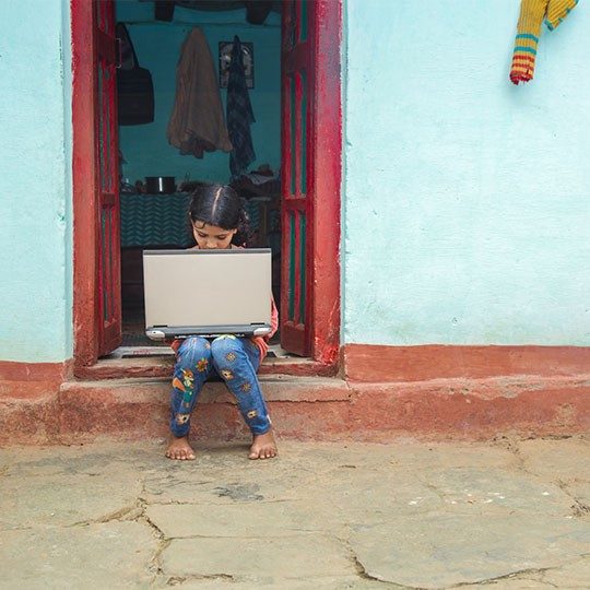 A child sitting in a doorway using a laptop connected to the internet by Viasat Community Internet