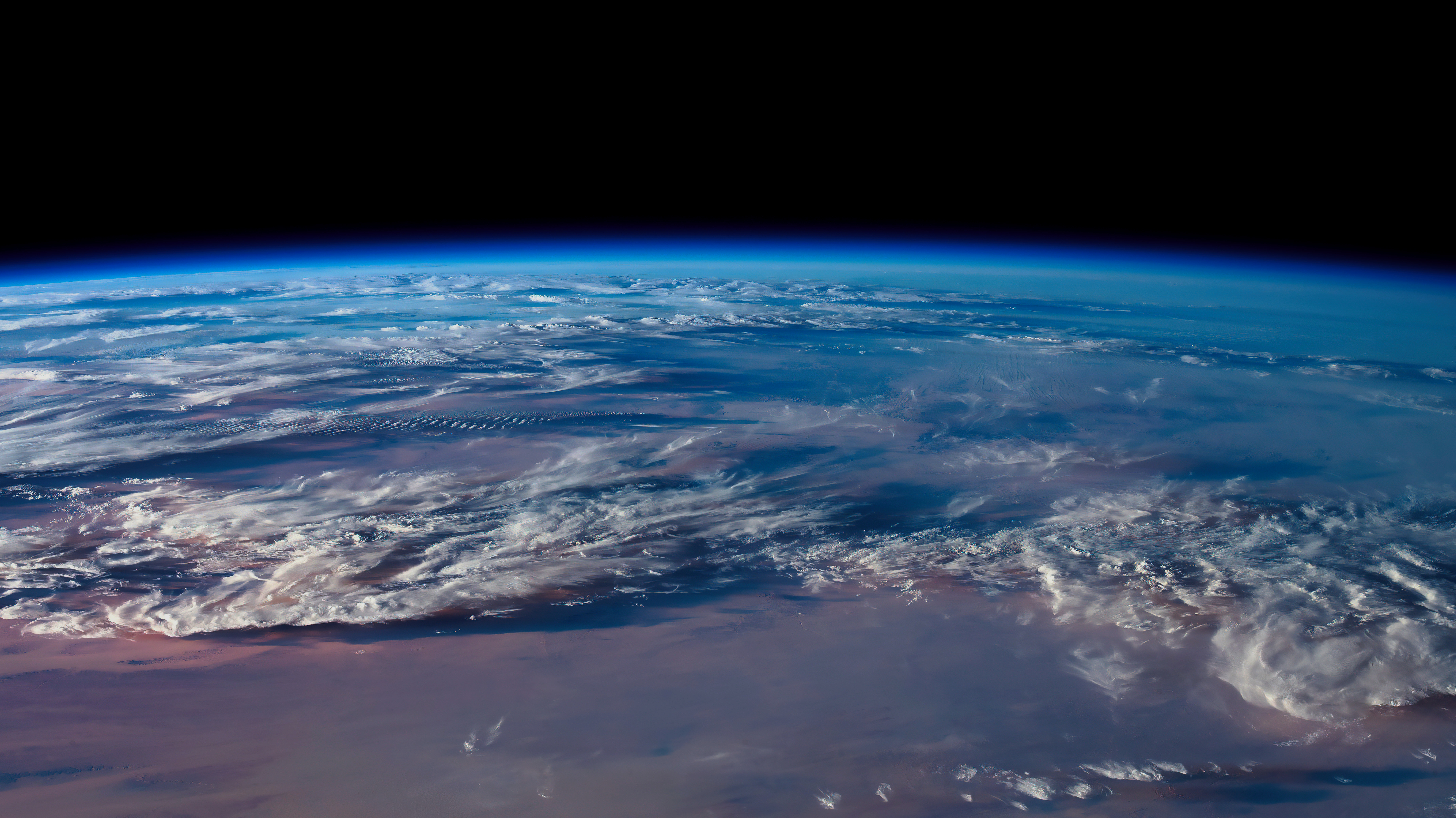 A view from space of a vast expanse of Earth, mostly covered by clouds, against the blackness of outer space. 