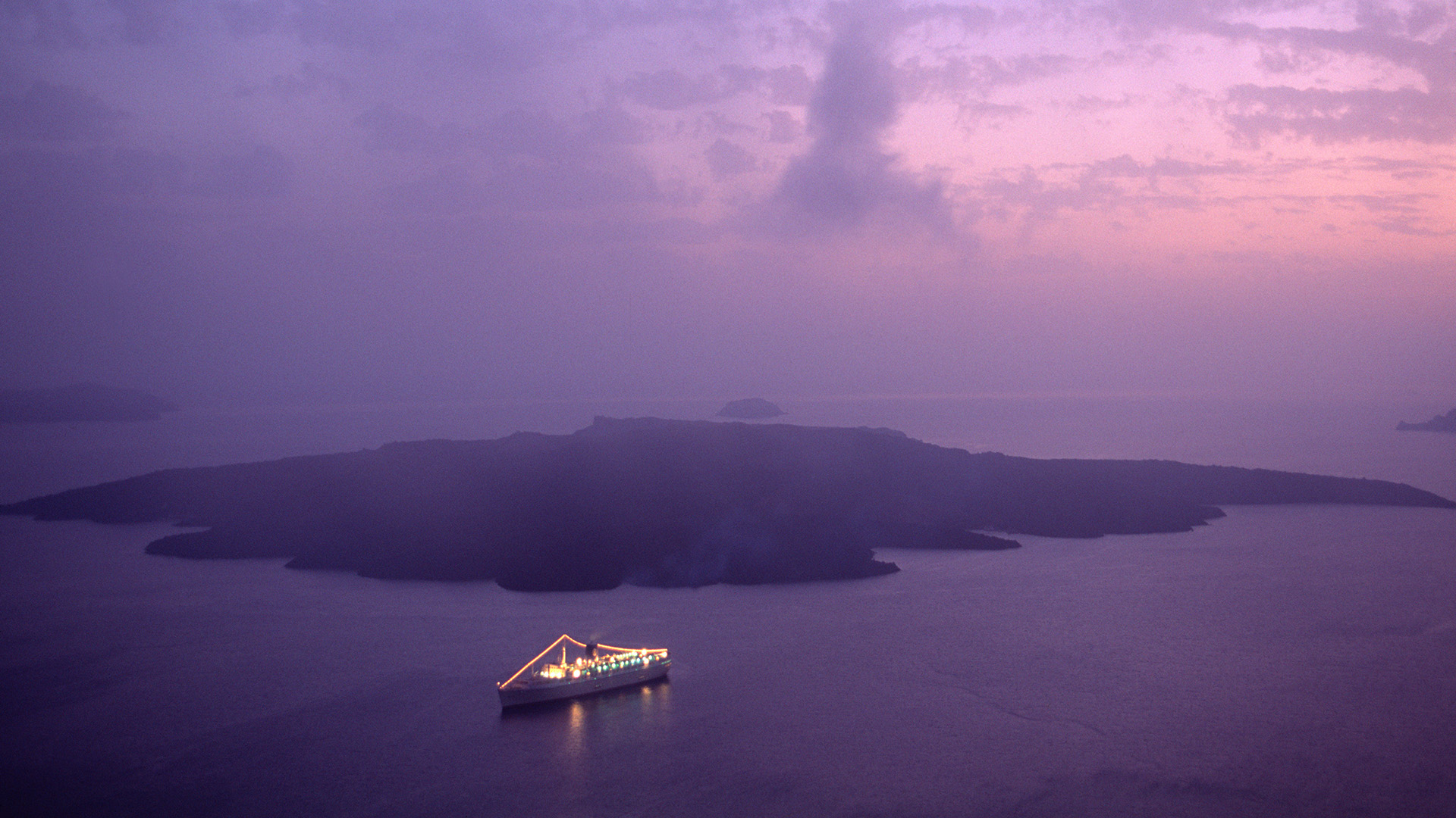An aerial view of a cruise ship with lights on deck and on the rigging, on the sea in front of a remote island.