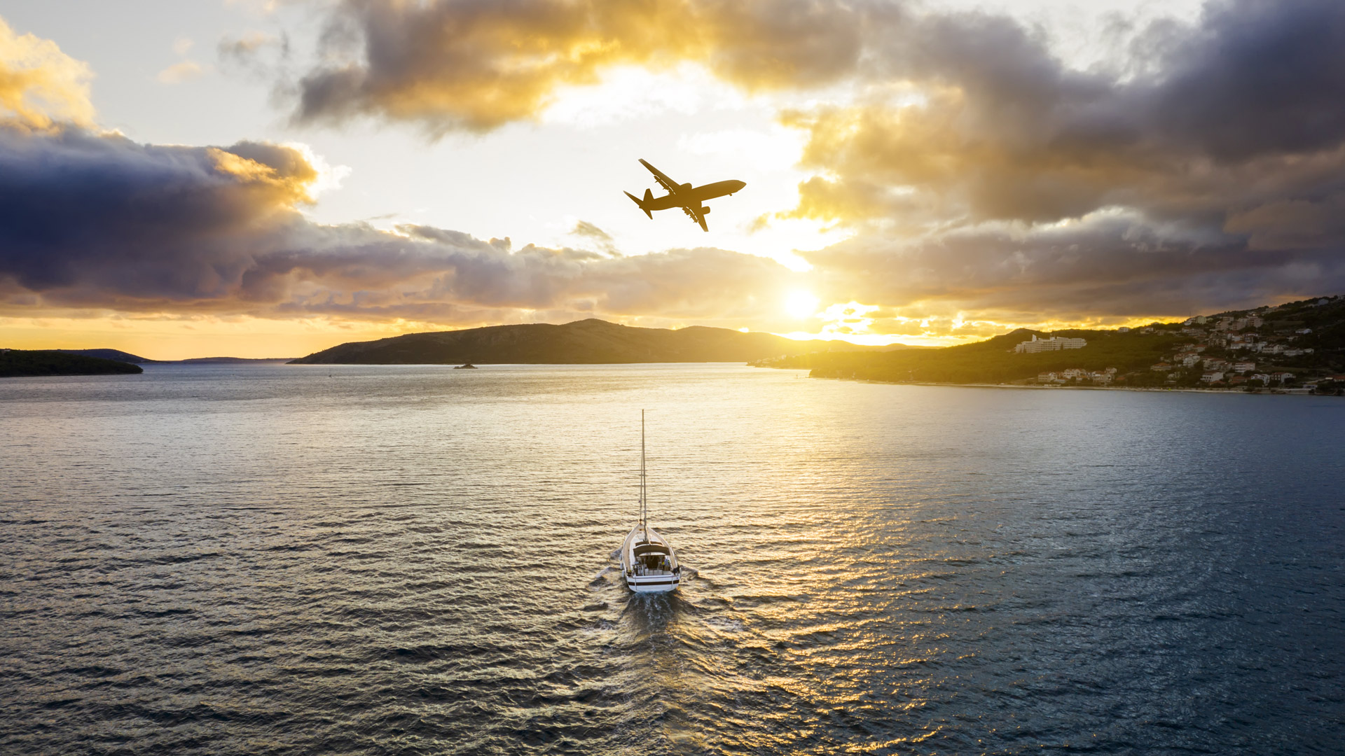 An airliner flies above a sailboat that’s alone on the sea at sunset, near some islands.