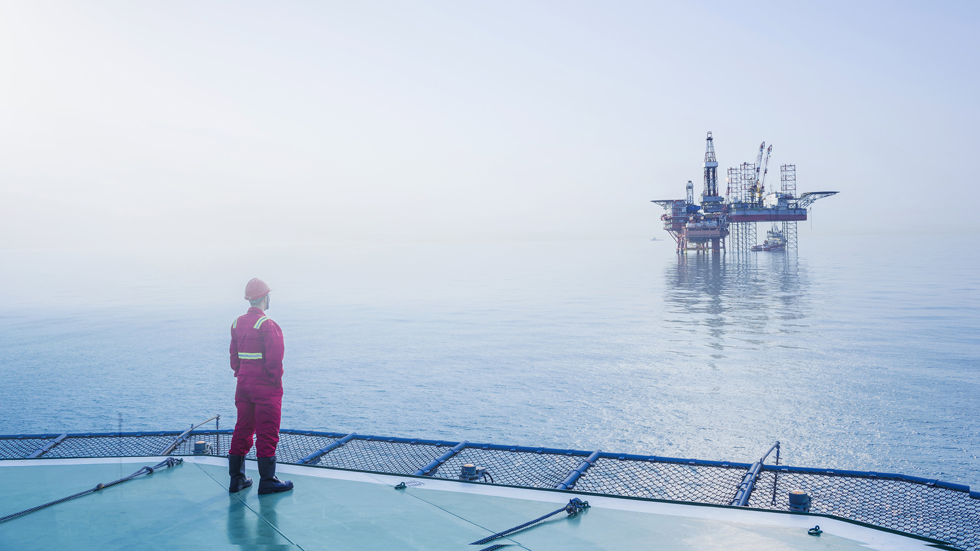 A person in a hard hat and work clothes stands on a platform in the ocean looking across the water to an oil-rig platform.