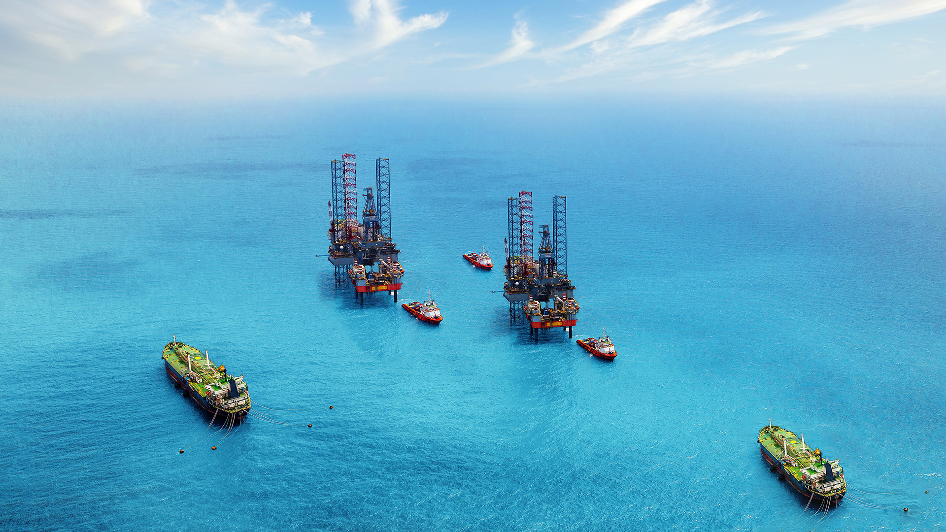 Two offshore oil rigs sit side by side, with boats nearby in a bright blue sea.