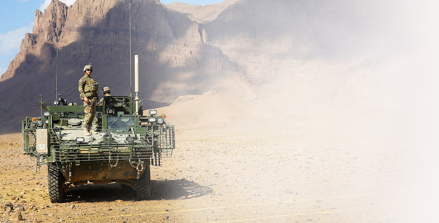 Military man wearing combat gear standing on the front of a green army vehicle against a rocky mountain backdrop