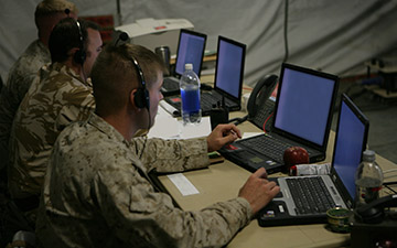 Three male warfighters utilizing satcom to communicate over their headsets, looking at laptops in a tactical operations center