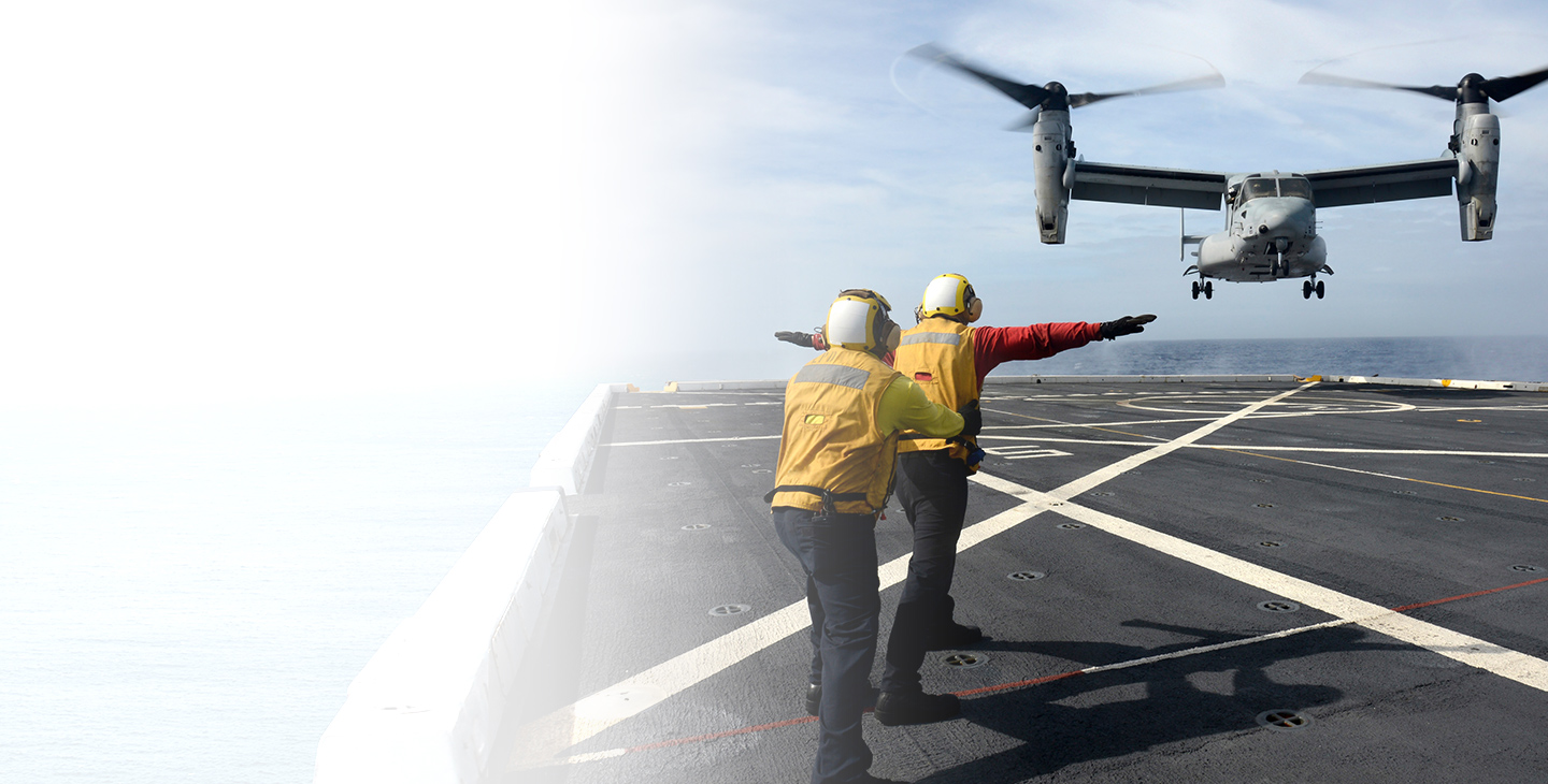 Two people using military satcom to communicate with a MV-22 landing on an aircraft carrier at sea