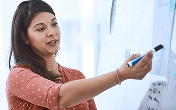 Dark-haired woman wearing a mauve polkadot shirt, holding an Expo marker training a defense support team