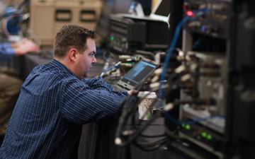 Brown haired man wearing a blue plaid shirt looking at an equipment screen in a server room