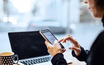 Woman wearing a black shirt, sitting behind a laptop while navigating on her smartphone