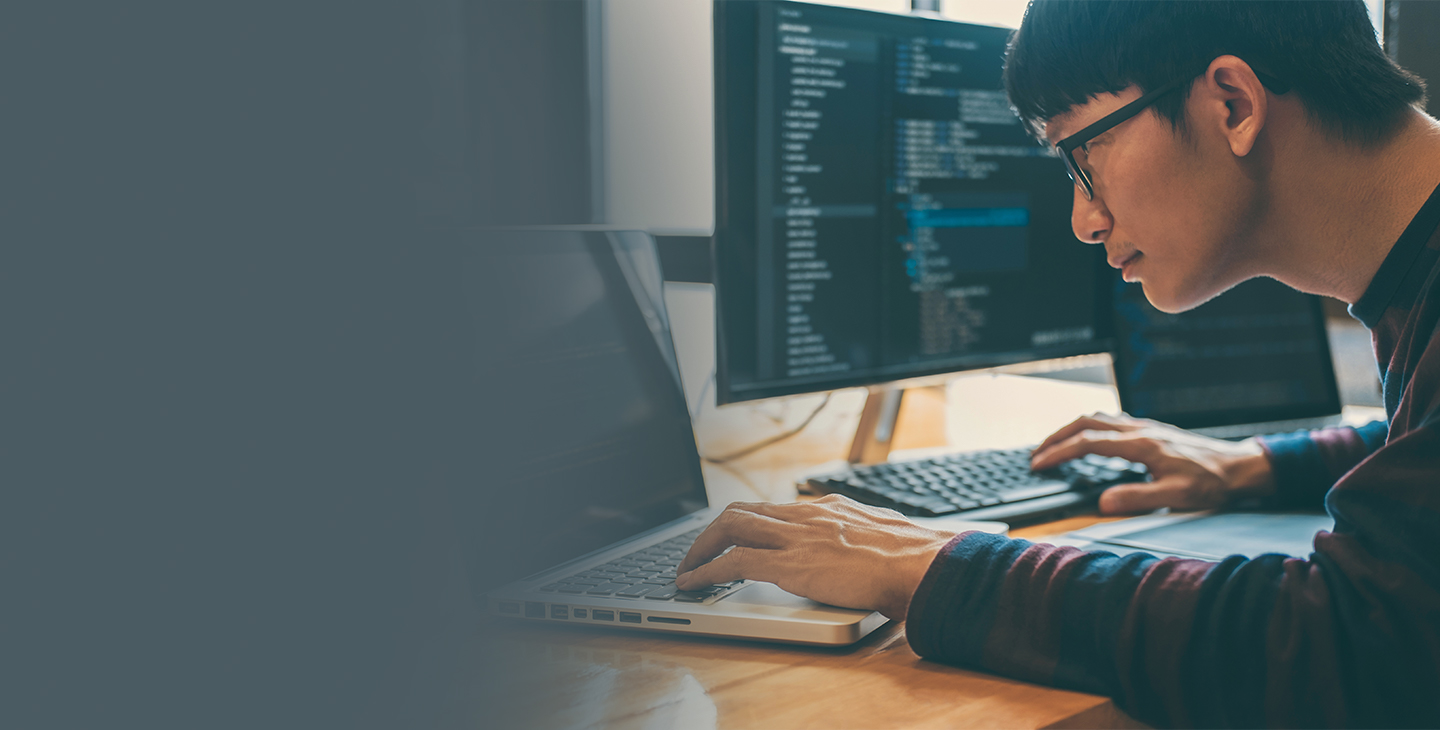 Man wearing glasses and a blue and maroon striped shirt working on cybersecurity protection on a laptop and desktop computer simultaneously