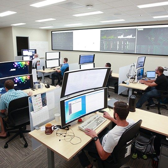 Team of six Viasat employees seated at desks with 2 large, stacked monitors working on cyber security operations