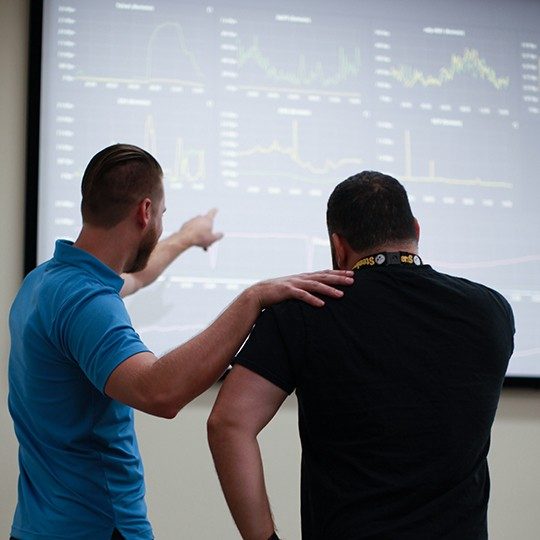 View of the backs of two men talking in a cyber SOC, one pointing at a large screen on the wall with multiple graphs