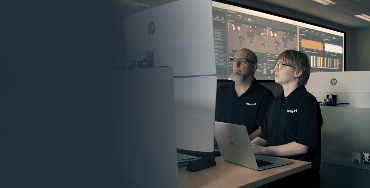 Two male Viasat employees wearing black polos featuring a white Viasat logo working in a cybersecurity operations center