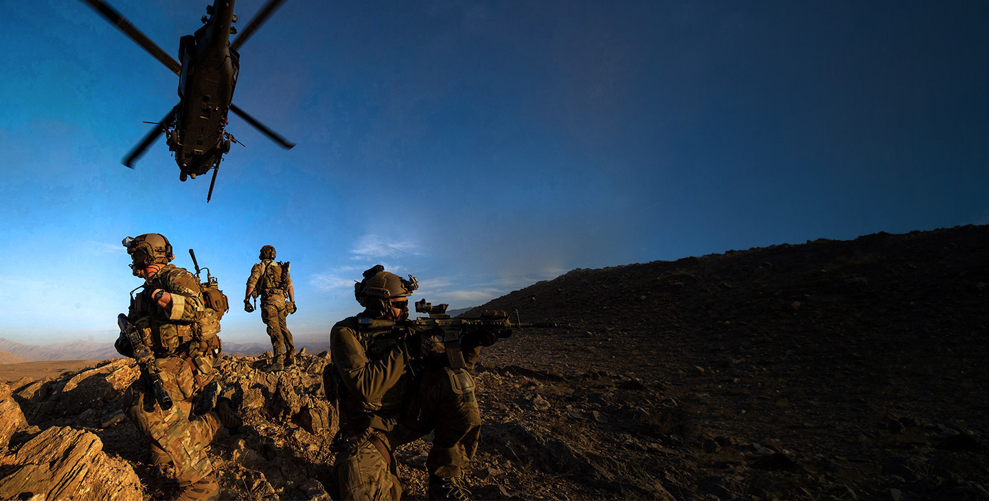 3 military men dressed in combat gear on watch on top of a rocky hill with a helicopter flying overhead