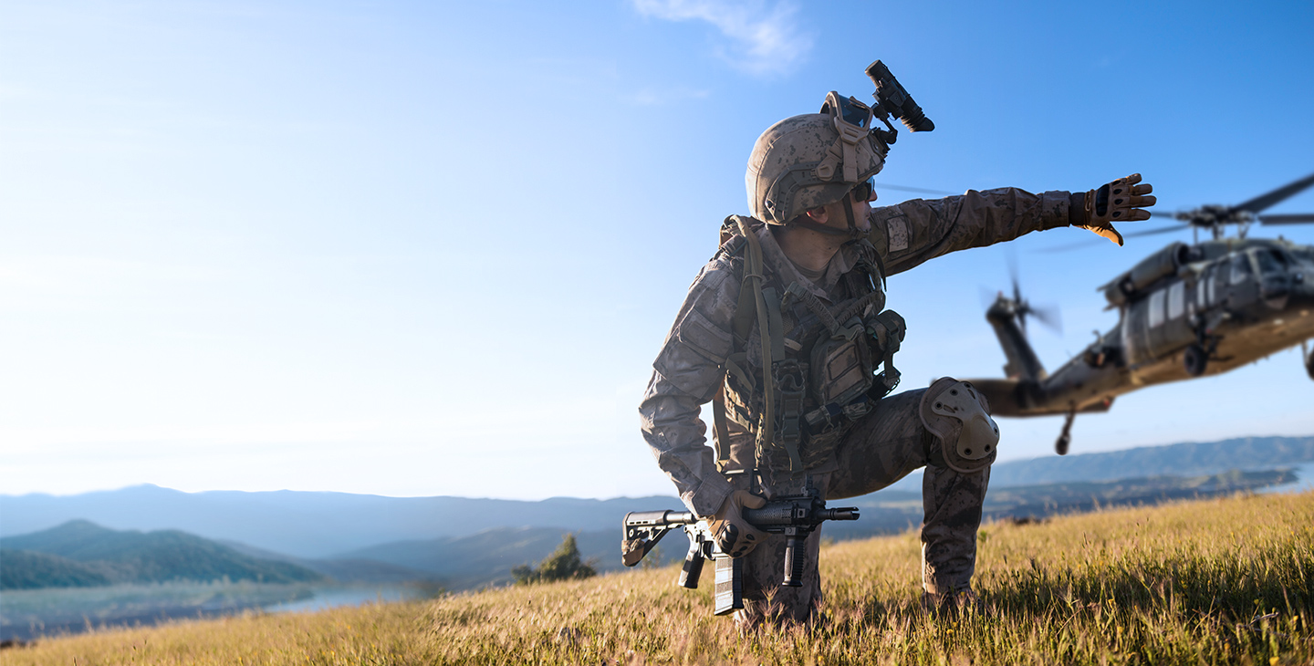 Military man dressed in a combat uniform, kneeling on ground holding a weapon and signaling to a helicopter
