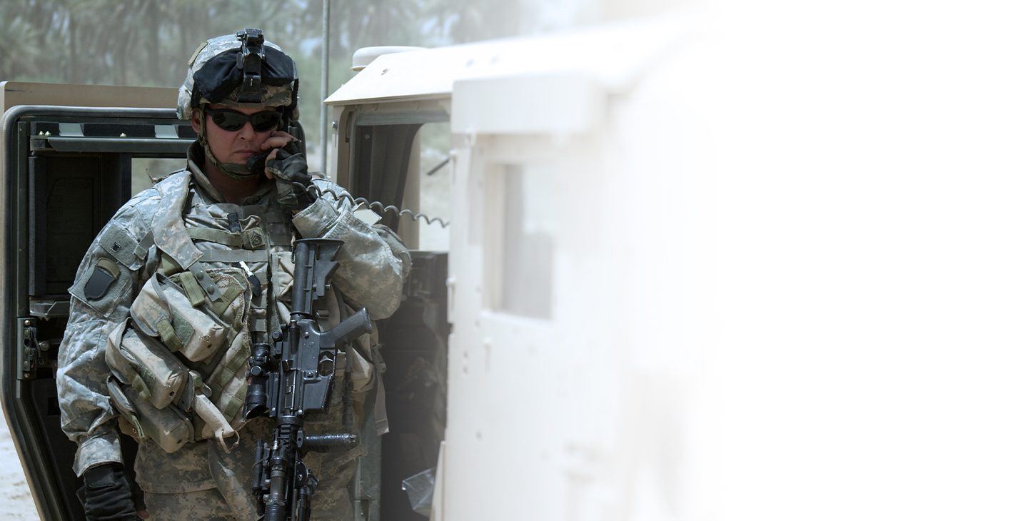 Man dressed in combat gear and wearing an military rifle, talking on a radio extending from a humvee