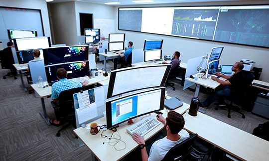 Seven men sitting at desks with large computer monitors analyzing data