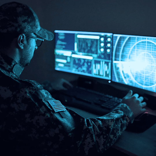 Man in camo uniform and cap wearing glasses sittng in the dark looking at two computer monitors