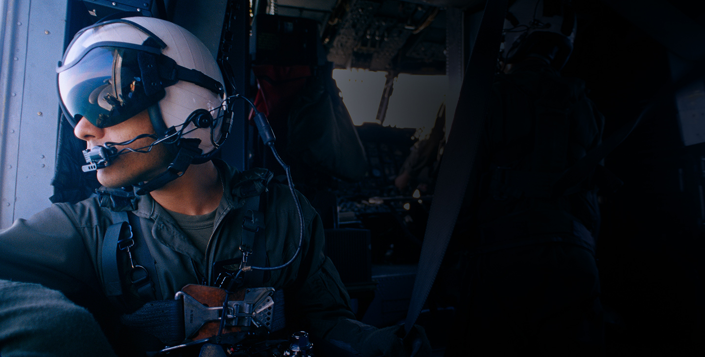 A male warfighter wearing a helmet, eye shield and headset looking out the side of a helicopter