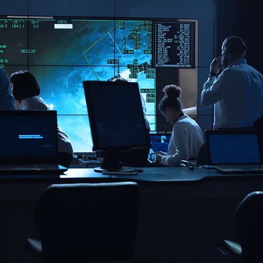 Two women and a man wearing a headset looking at a large screen in a control room