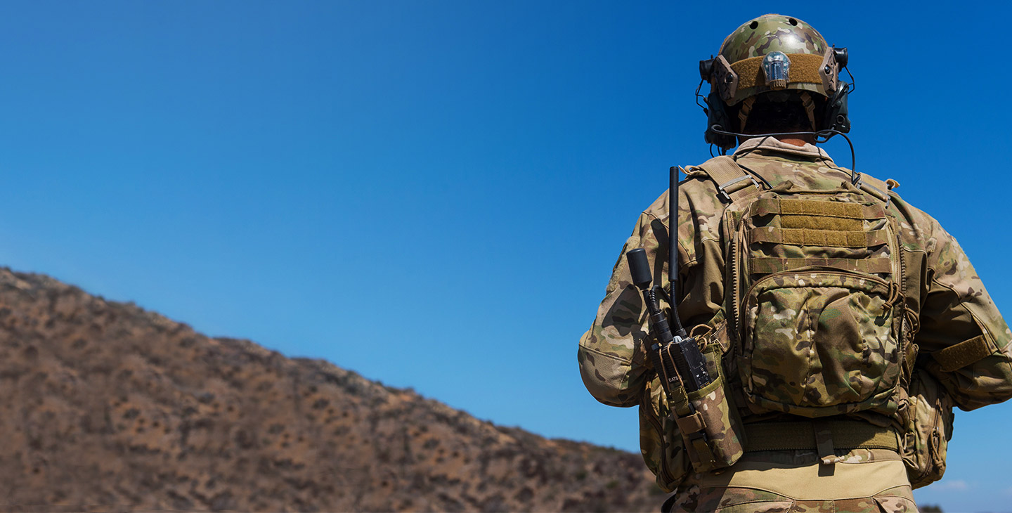 View of the back of a soldier in the field, wearing a backpack holding a BATS-D handheld radio with antennas attached