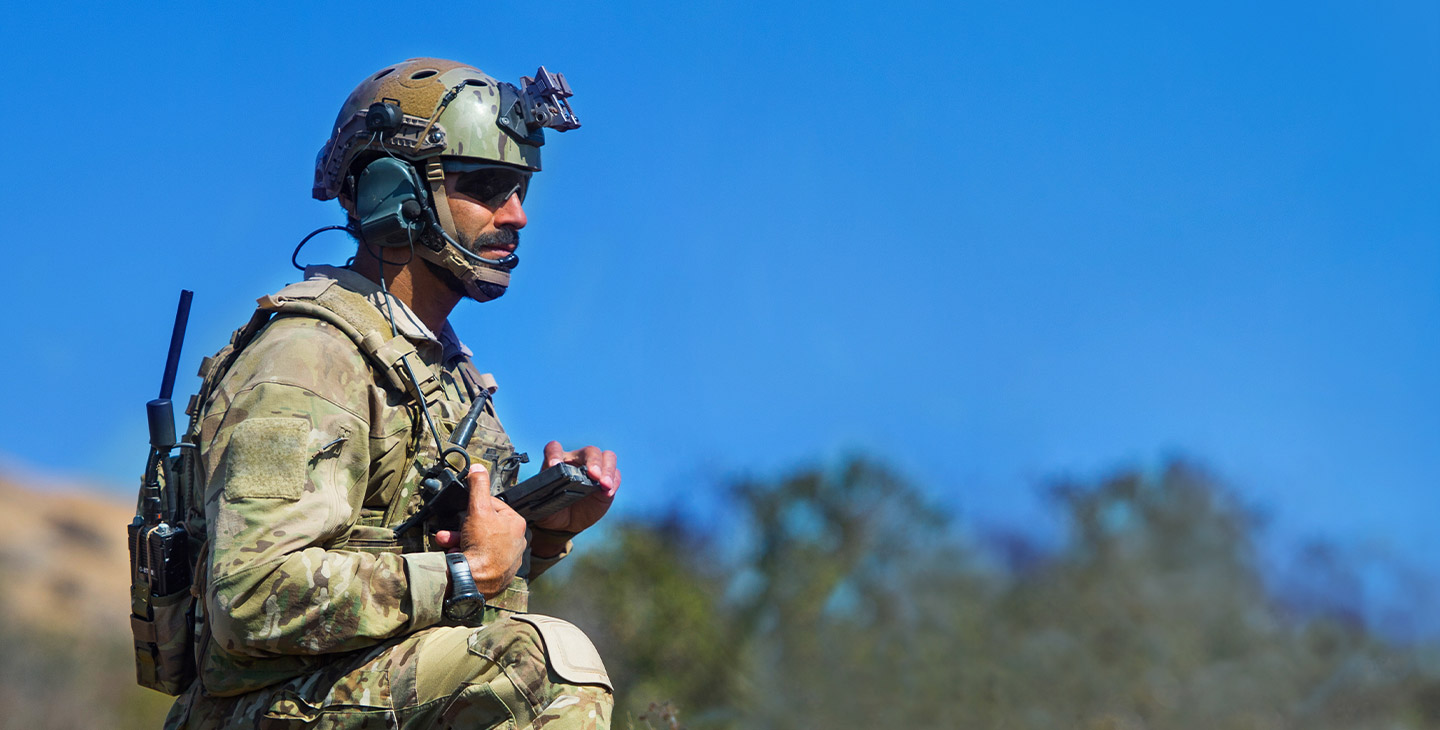 Close-up of a soldier on one knee in the field, wearing army gear and a backpack holding a BATS-D AN/PRC-161 handheld radio with antennas attached