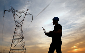 Technical engineer wearing a hard hat and holding a latptop looking at a telecommunications tower at dusk