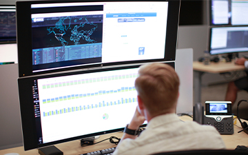 Male Viasat employee sitting at a desk in front of two stacked monitors in a cybersecurity operations center